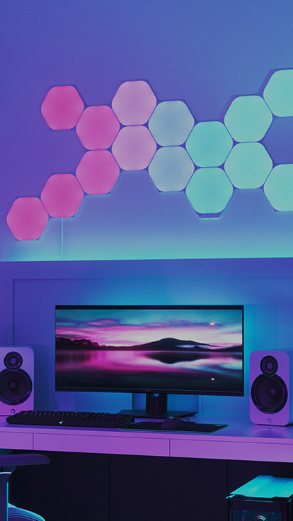 Esta es una imagen de Nanoleaf Shapes Hexagons en una pared encima del monitor de un escritorio. Son luces RGB perfectas para los jugadores en el entorno de tu hogar. Los paneles de luz modulares que cambian de color se conectan entre sí con conectores y tienen más de 16 millones de colores.