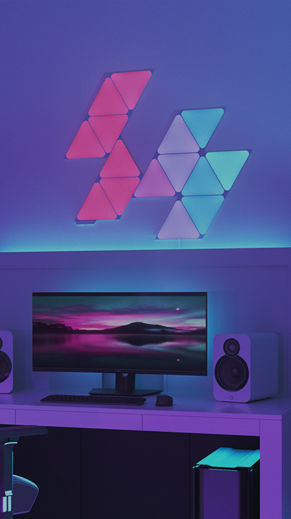 Esta es una imagen de un diseño de 15 paneles de Nanoleaf Shapes Triangles montados en la pared sobre un escritorio y detrás del monitor de una estación de batalla de juegos. Estas luces RGB tienen más de 16 millones de colores y son perfectas para los jugadores en tu casa.