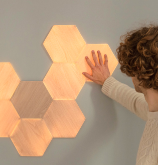 Esta es una imagen de Nanoleaf Elements Wood Look Hexagons montados en una pared. Los paneles de luz inteligentes reaccionan al tacto y a la música, y llenan tu hogar con una iluminación suave. Toca los paneles con apariencia de madera o reproduce tus canciones favoritas para encender tu espacio con un brillo dinámico.